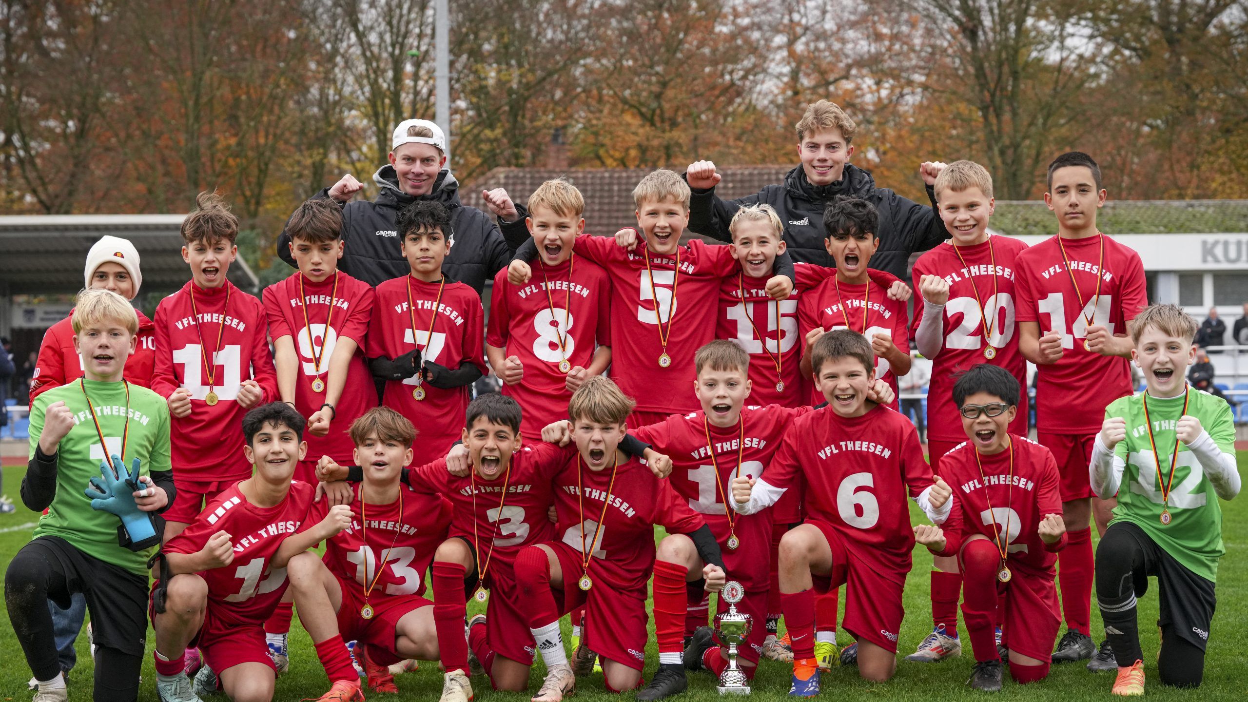 Luftaufnahme des Sportkomplexes des VfL Theesen mit einem Fußballplatz, umgeben von Feldern und Wohnhäusern, in einer grünen Landschaft. Im Vordergrund ist ein Kunstrasenplatz mit Flutlichtanlagen zu sehen, im Hintergrund liegt die Stadt Bielefeld.