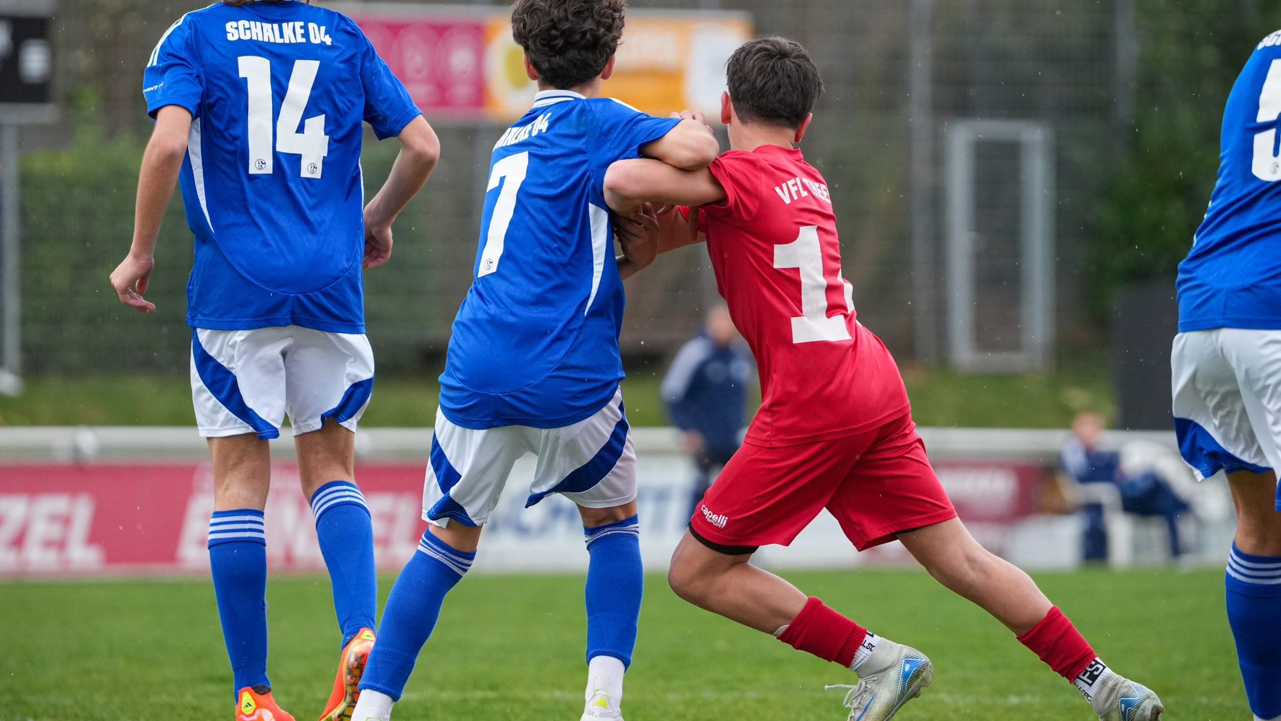Luftaufnahme des Sportkomplexes des VfL Theesen mit einem Fußballplatz, umgeben von Feldern und Wohnhäusern, in einer grünen Landschaft. Im Vordergrund ist ein Kunstrasenplatz mit Flutlichtanlagen zu sehen, im Hintergrund liegt die Stadt Bielefeld.